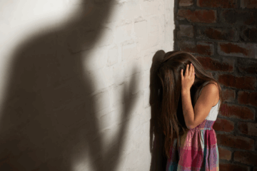 Une petite fille traumatisée baisse la tête et se couvre les oreilles face à l'ombre d'un adulte oppressant.