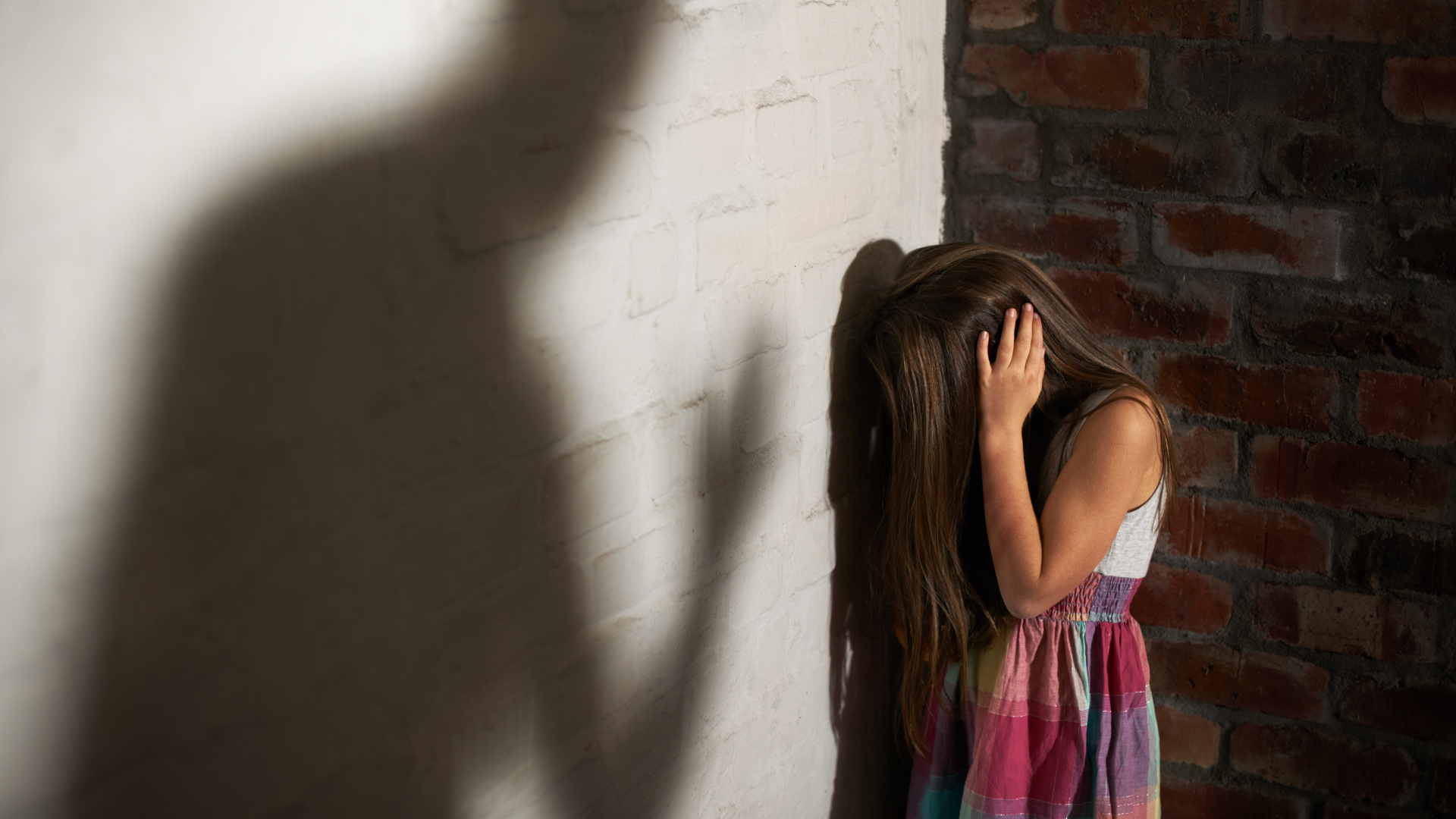 Une petite fille traumatisée baisse la tête et se couvre les oreilles face à l'ombre d'un adulte oppressant.